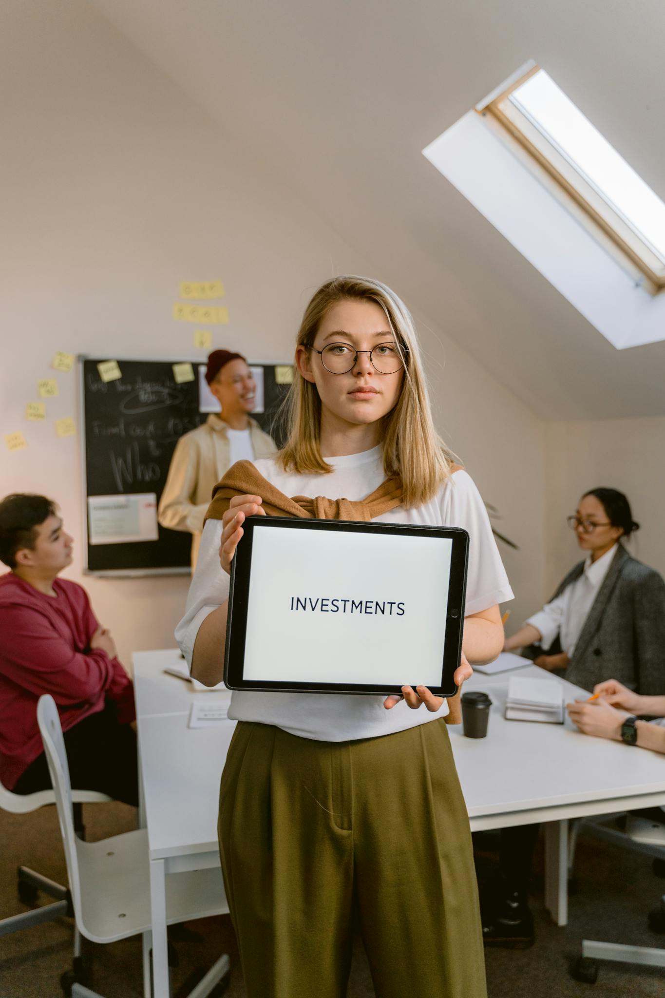 Young woman holding a tablet with 'INVESTMENTS' text in a modern office setting.